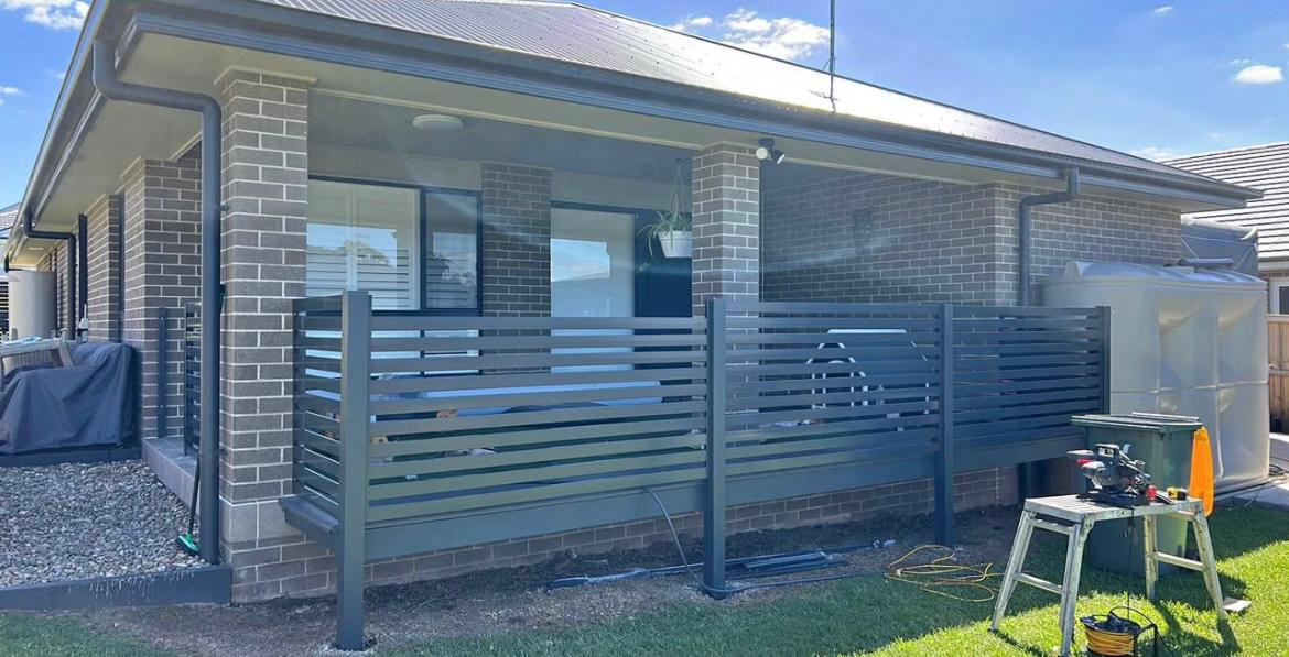 Grey slat privacy screen being fitted around a brick patio, with a work trestle on the lawn