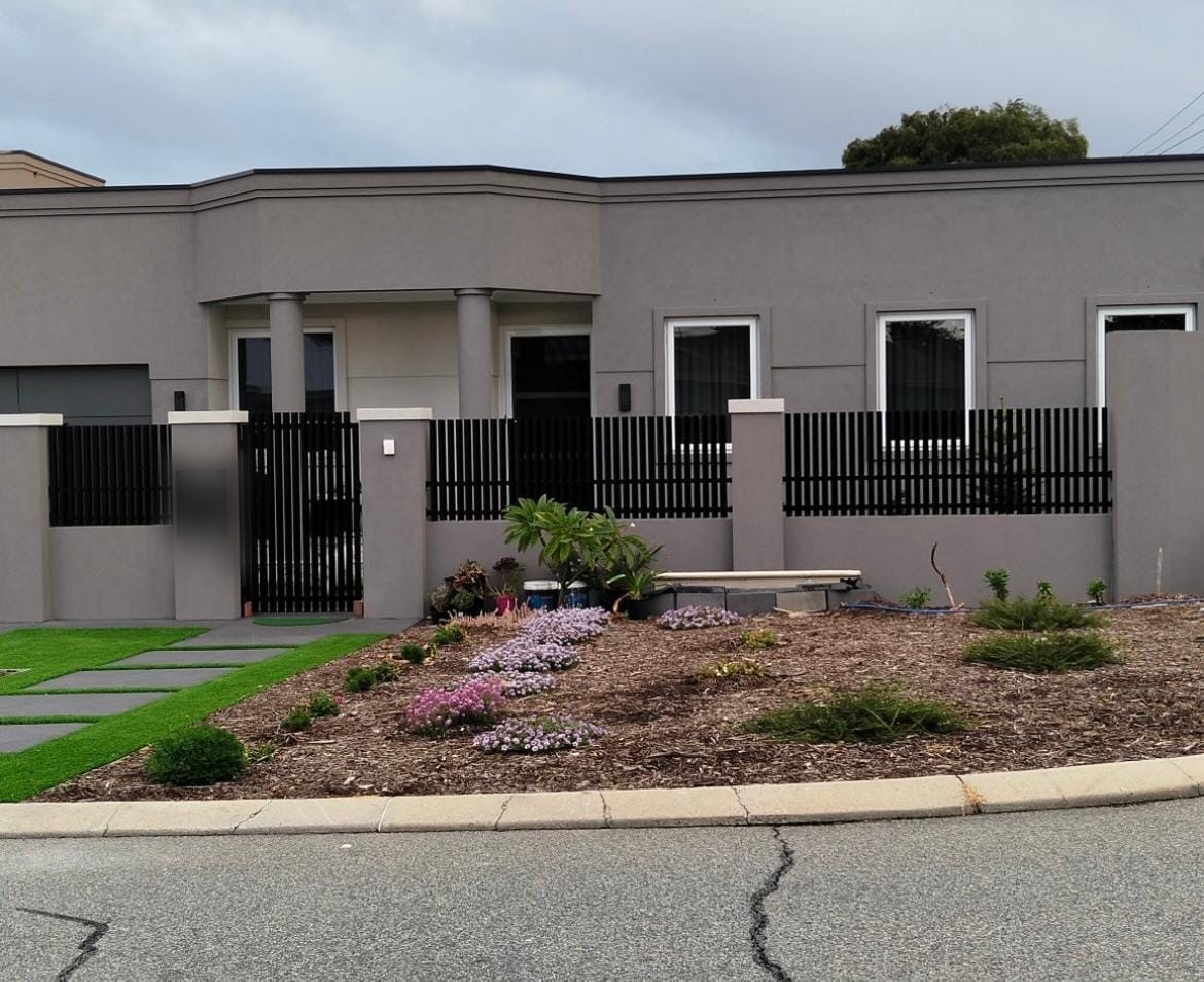 Black blade front fence and pedestrian gate between grey rendered pillars with a garden bed in front